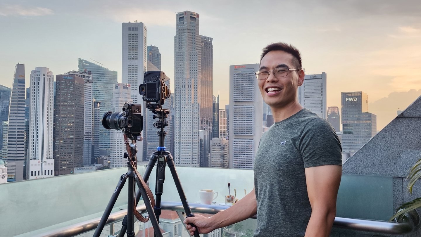 Henry Lam smiling beside two tripod-mounted cameras on a rooftop, the Singapore skyline behind him at the crossover hour